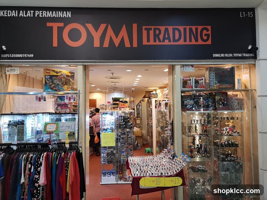 The exterior of a toy store called Toymi Trading. The storefront displays various toys and collectibles in glass cabinets and on shelves. There are also racks of clothing visible. The shop's name and logo are prominently displayed on the facade.