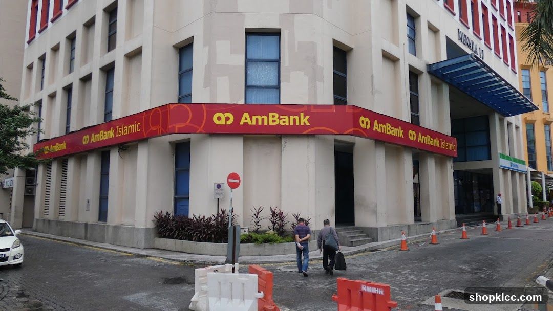 A corner view of a modern building with a prominent red and yellow AmBank and AmBank Islamic signage wrapping around the facade. The building has cream-colored walls and multiple windows. Two men are walking away from the entrance. Orange traffic cones and white barriers are visible on the street in front of the building.