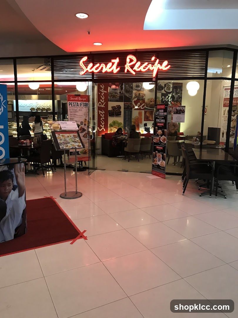 The exterior of a Secret Recipe restaurant with a prominent neon sign. Inside, tables and chairs are visible, along with some patrons. There are also promotional posters and a menu stand outside the entrance.