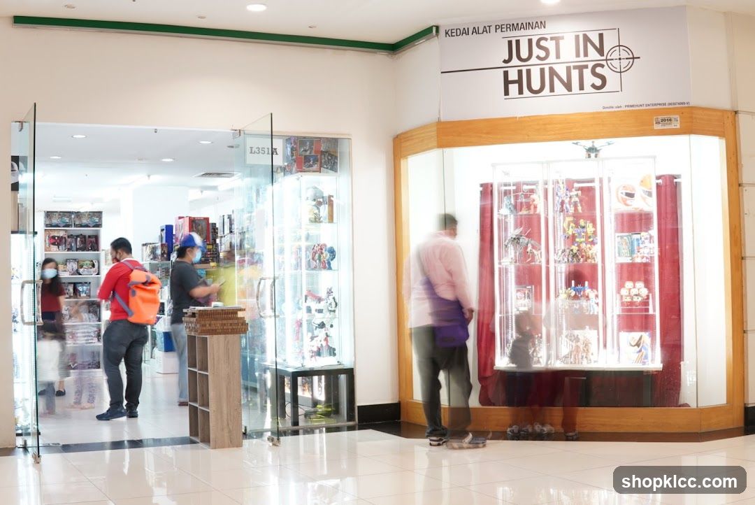 The exterior of a toy store with a glass entrance and display cases. The store's name, "JUST IN HUNTS," is visible on a sign above the entrance. Inside, shelves are stocked with various toys and collectibles. People are seen browsing the merchandise.