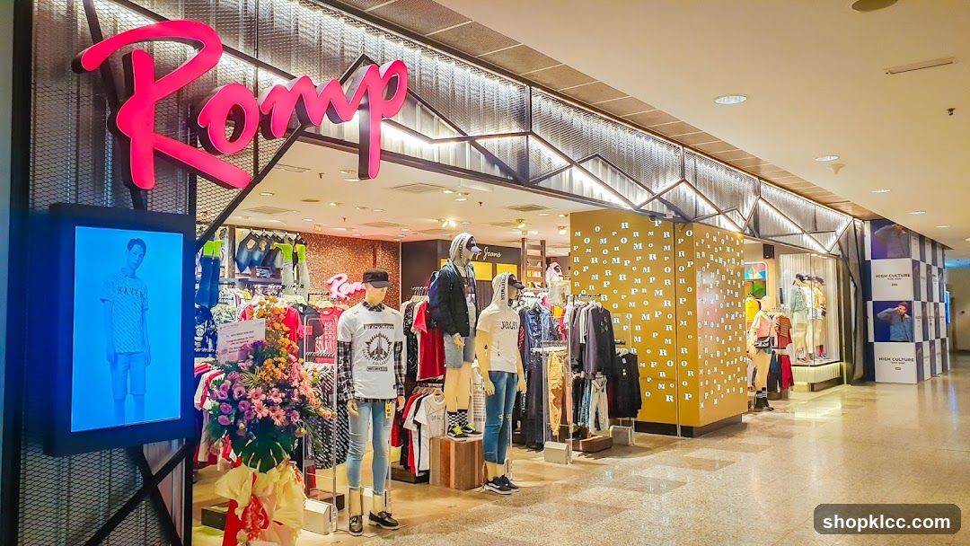 The exterior of a clothing store with a large pink "Romp" sign above the entrance. Mannequins are displayed in the window and inside the store, showcasing various apparel. A digital display shows a model wearing a t-shirt and shorts. The store features a modern design with metallic accents and patterned walls.