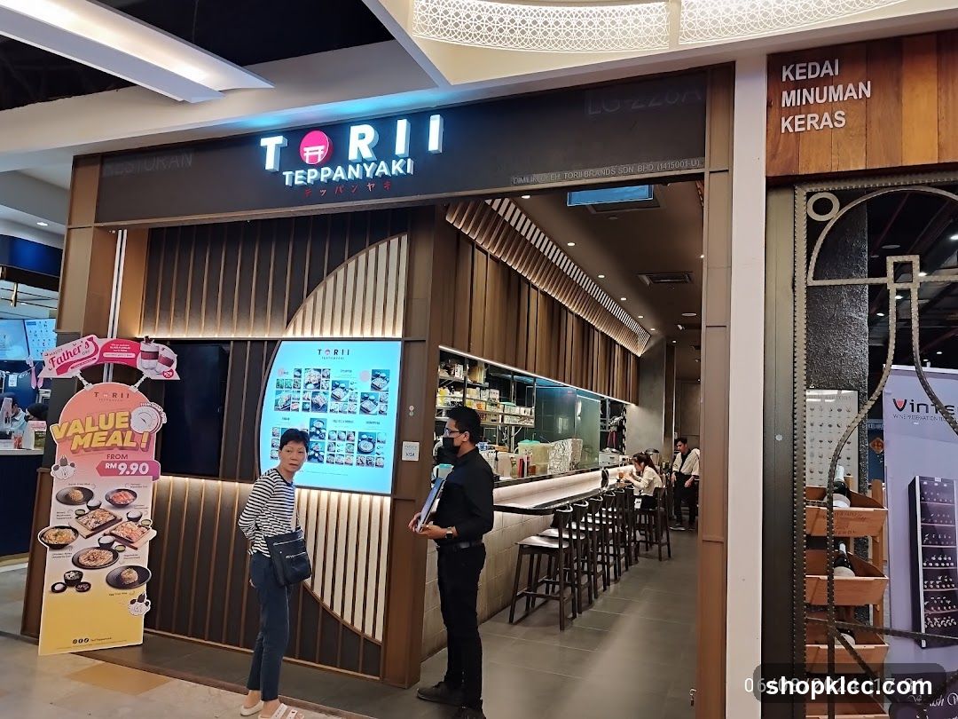 The exterior of a Torii Teppanyaki restaurant. A large sign above the entrance reads "TORII TEPPANYAKI". To the left, a promotional stand displays "VALUE MEAL! FROM RM 9.90" with images of food. Inside, a counter with stools and diners can be seen.