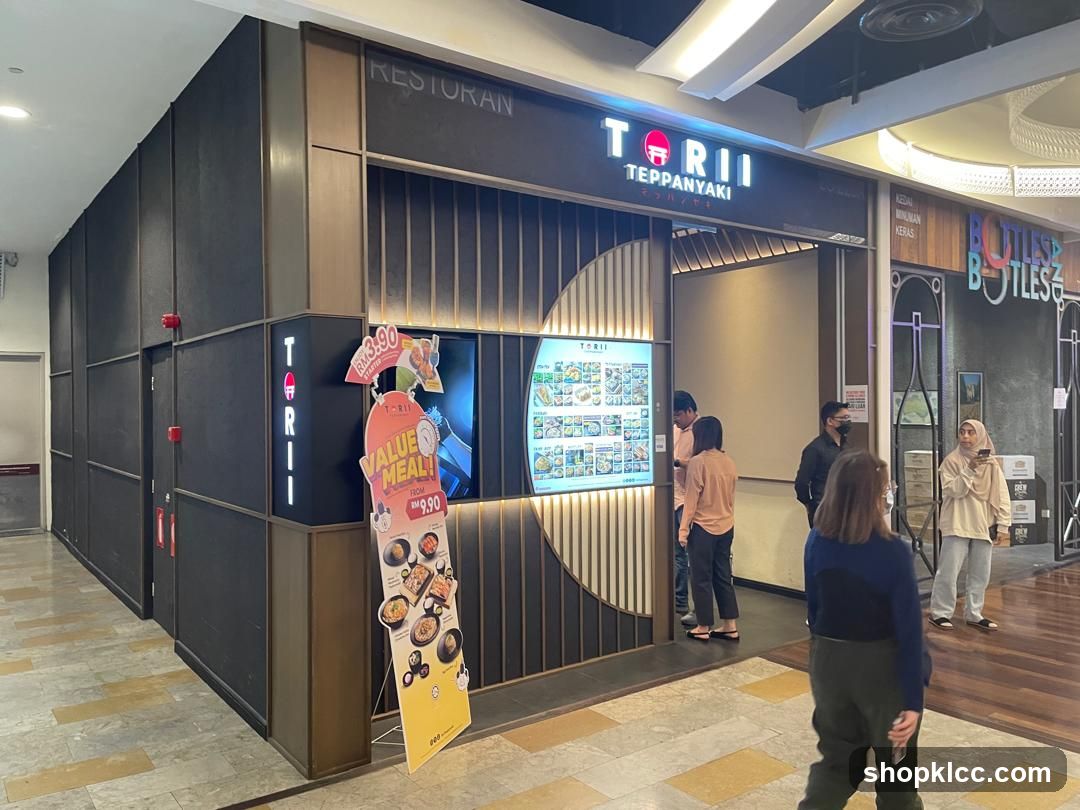 The exterior of a Torii Teppanyaki restaurant. The storefront features a dark facade with vertical wooden slats and a prominent sign with the restaurant's name and logo. A digital menu board is visible to the left of the entrance, displaying various food items. A promotional stand with a "Value Meal" offer is placed in front of the entrance. Several people are seen near the entrance, some looking at the menu.