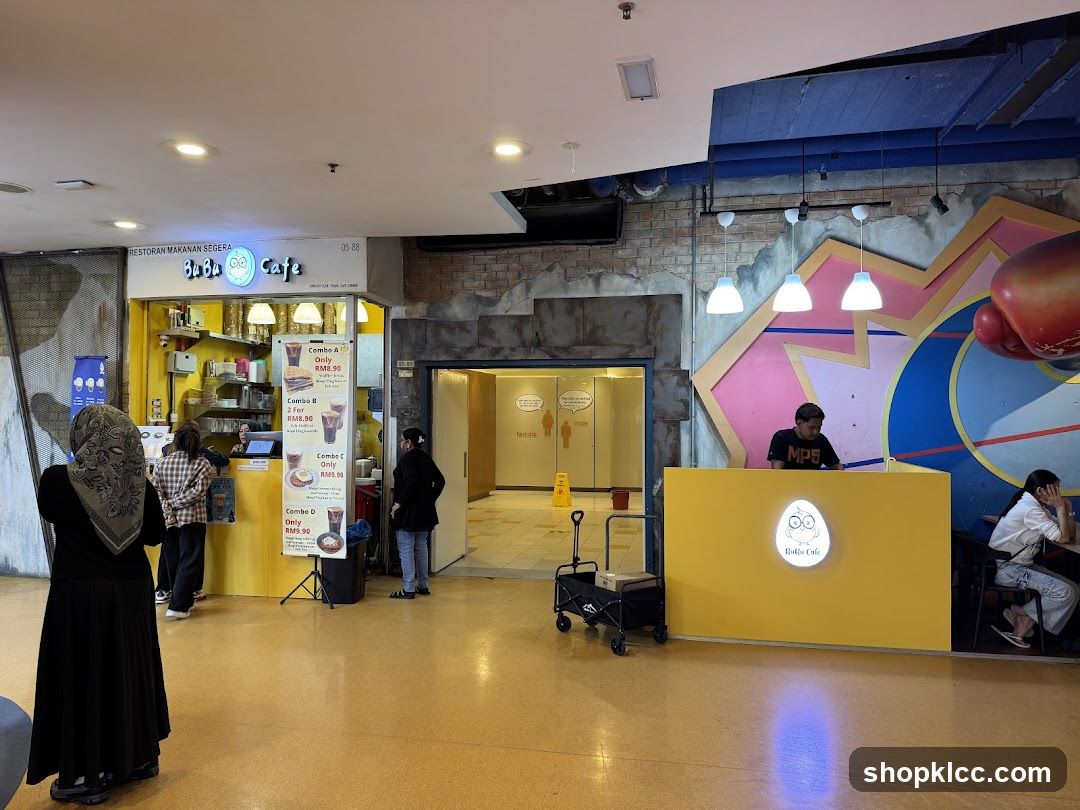 A bright yellow cafe counter with the Bubu Cafe logo is visible. Behind the counter, a person is working. To the left, a food stall with a similar yellow facade displays a menu with various combos. People are seen queuing and ordering. The cafe is located in an indoor area with a distinctive colorful wall design and overhead lighting.