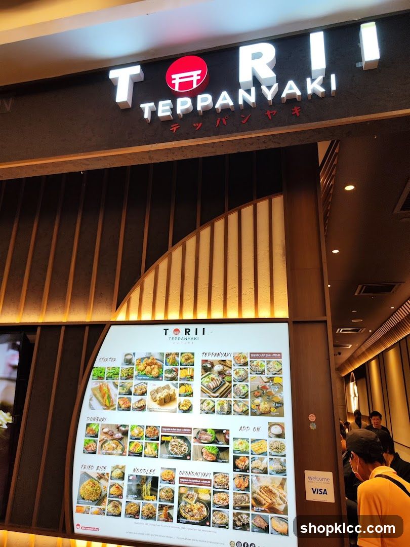 The exterior of a restaurant with a large illuminated sign that reads "TORII TEPPANYAKI". Below the sign is a large menu board displaying various food items with images. The restaurant has a modern design with wooden accents.