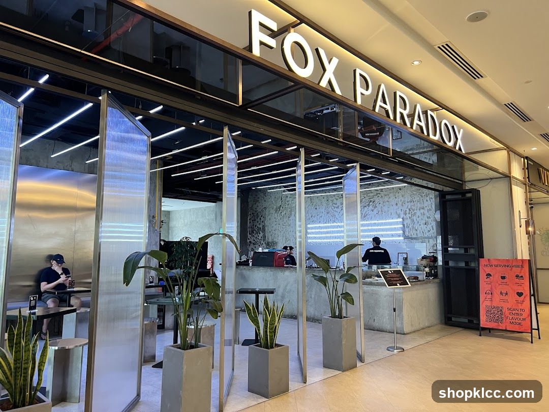 The exterior of a modern cafe with large glass panels and a prominent illuminated sign that reads "FOX PARADOX". Inside, a counter area is visible with staff members. Several potted plants are placed in front of the cafe. A person is seated at a table inside.