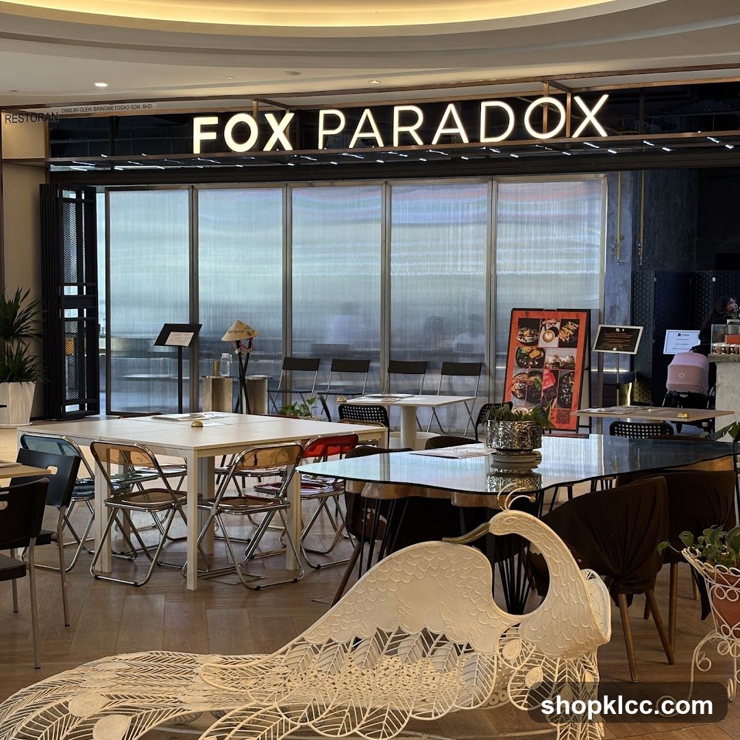 The exterior of a restaurant called Fox Paradox. The restaurant has a modern design with large glass windows and a prominent neon sign. Inside, tables and chairs are set up for dining. A decorative peacock chair is visible in the foreground.