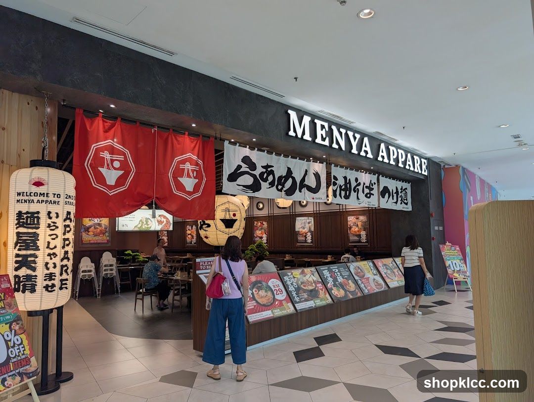 The exterior of a ramen shop called Menya Appare. Large white lanterns with Japanese text and a shop logo are displayed outside. Red banners with the shop's logo hang above the entrance. The shop's name is prominently displayed in large white letters above the entrance. Several display boards with images of food and prices are arranged in front of the shop.