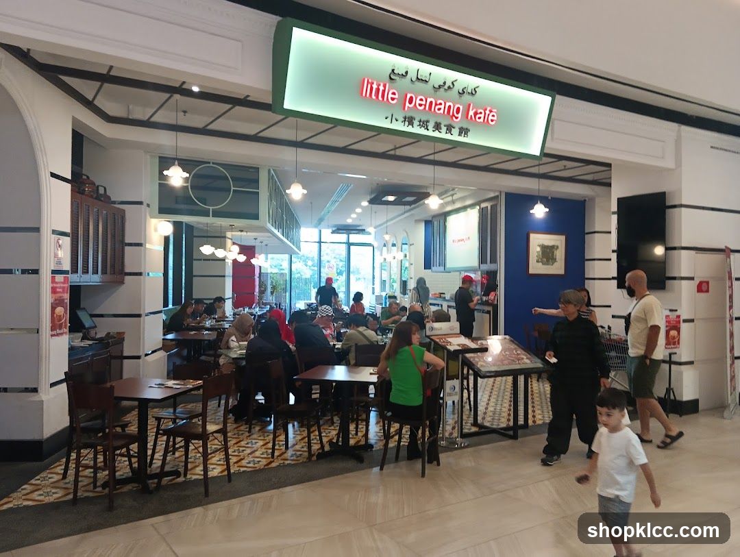 The exterior of a restaurant called Little Penang Kafe. The restaurant has a bright green sign with the name in English and Chinese. Inside, tables and chairs are set up for dining, and customers are seated.