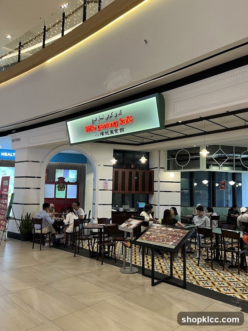 The exterior of a restaurant called Little Penang Kafe, with its name displayed in English, Arabic, and Chinese. The restaurant has an open-air seating area with tables and chairs. Several patrons are seated at the tables, some eating and others conversing. A menu stand is placed in front of the restaurant, displaying various food items. The restaurant is located inside a mall, with other shops visible in the background.