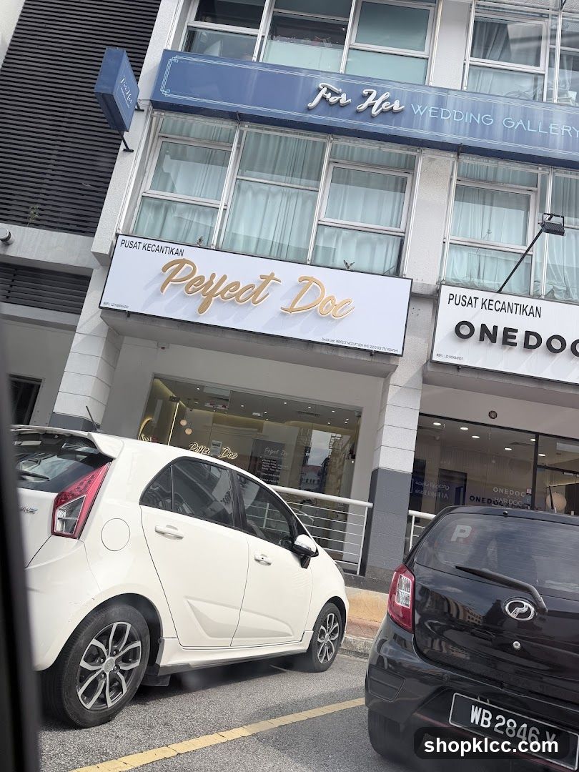 A storefront for a wedding gallery and beauty center. The building has multiple floors with large windows. The main sign reads "Perfect Doc" in gold lettering, with "PUSAT KECANTIKAN" above it. Another sign on the upper floor says "For Her WEDDING GALLERY". A car is parked in front of the building.