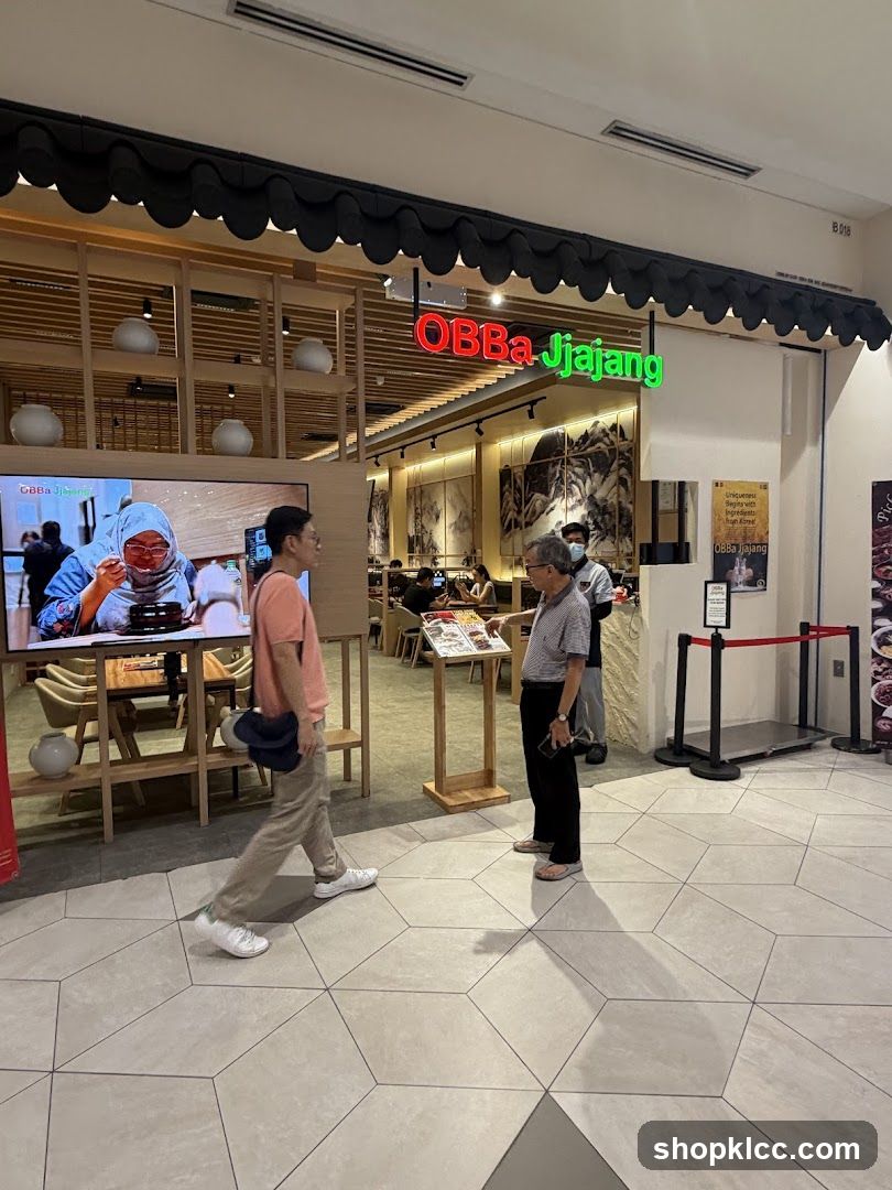 The exterior of a restaurant with a prominent neon sign that reads "OBBa Jjajang". The restaurant has a modern interior with wooden accents and decorative vases on shelves. A large screen displays an image of someone eating. Two men are standing near the entrance, one looking at a menu on a stand.