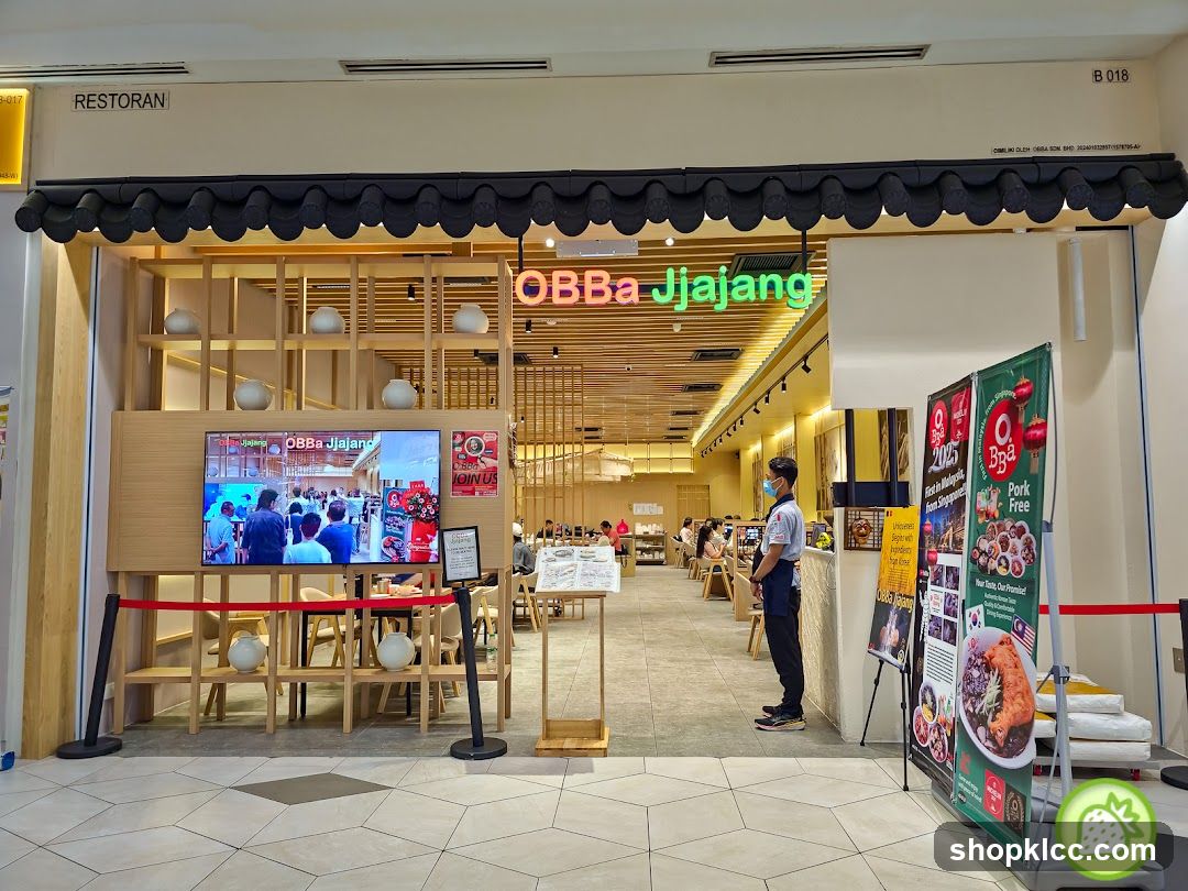 The exterior of a restaurant with the name OBBa Jjajang prominently displayed in neon green letters above the entrance. A large screen displays images of people and food. To the right, promotional banners advertise "2025 First in Malaysia from Singapore" and "Pork Free". The restaurant interior is visible, with tables and chairs set up for dining.
