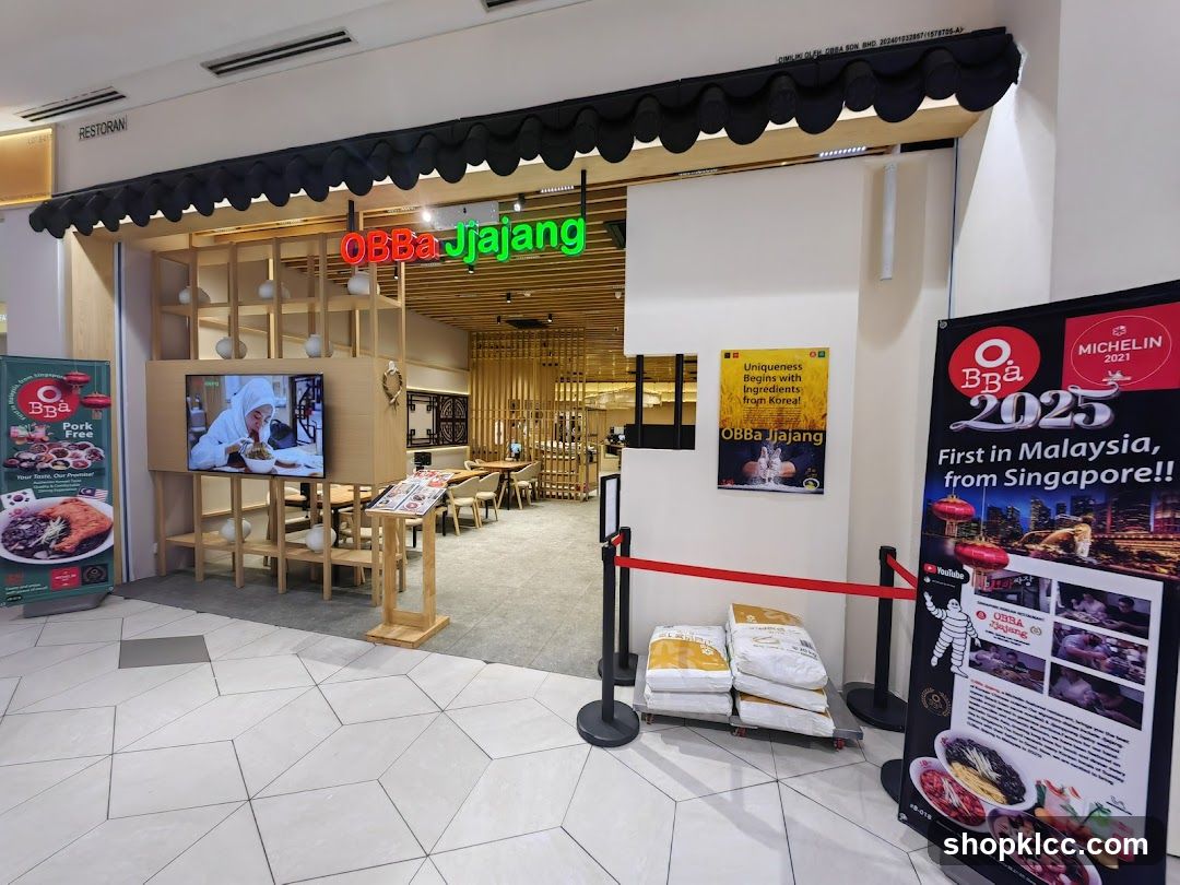 The exterior of a restaurant called OBBa Jjajang. The entrance features a wooden facade with shelves and a television screen displaying food. A black awning with decorative scalloped edges is above the entrance. To the left, a poster advertises "Pork Free" dishes. To the right, a large banner announces "MICHELIN 2021" and "2025 First in Malaysia, from Singapore!!". Red velvet ropes guide visitors.