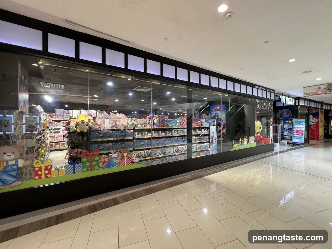 A wide shot of a toy store called Toy World, viewed from a mall concourse. The store has a large glass facade displaying shelves filled with various toys, including stuffed animals, action figures, and board games. Decorative elements like cartoon characters and gift boxes are visible along the bottom of the window. To the right, a smaller adjacent store is also visible, with a sign that reads "TOY WORLD". A promotional poster is displayed outside this second store.