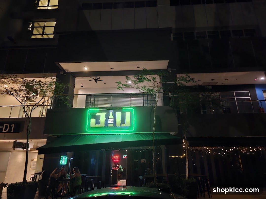 A neon green sign with the text "3JU" and Korean characters is illuminated above the entrance of a bar. The bar has a green awning and outdoor seating with people visible. The building has multiple floors with windows.