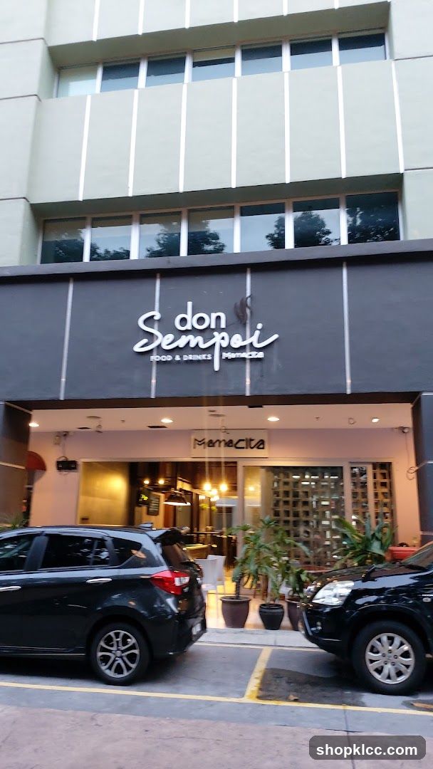 The exterior of a restaurant called Don Sempoi Food & Drinks. The name is displayed in a white, cursive font on a dark gray facade. Below this, the word "Mamacita" is visible on a smaller sign. The entrance is visible with glass doors and windows, and interior lighting can be seen. Two cars are parked in front of the establishment.