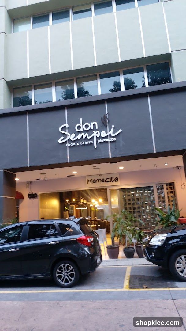 The exterior of a restaurant with a dark gray facade. The restaurant name "don Sempoi" is displayed in white cursive lettering above "FOOD & DRINKS" in smaller white text. Below this, the word "Mamacita" is also visible. The entrance to the restaurant is open, revealing a warmly lit interior with tables and chairs. Two cars are parked in front of the establishment.