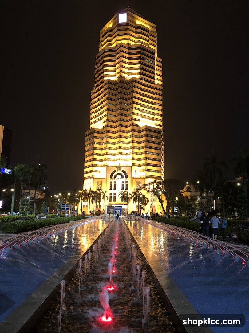 A tall, illuminated skyscraper at night, with a prominent arched entrance at its base. In the foreground, a long water fountain with red lights runs towards the building. Palm trees and other greenery line the walkway. Several people are visible near the entrance and along the fountain. The building has a distinctive stepped facade, and the words "PUBLIC BANK" are visible above the entrance.