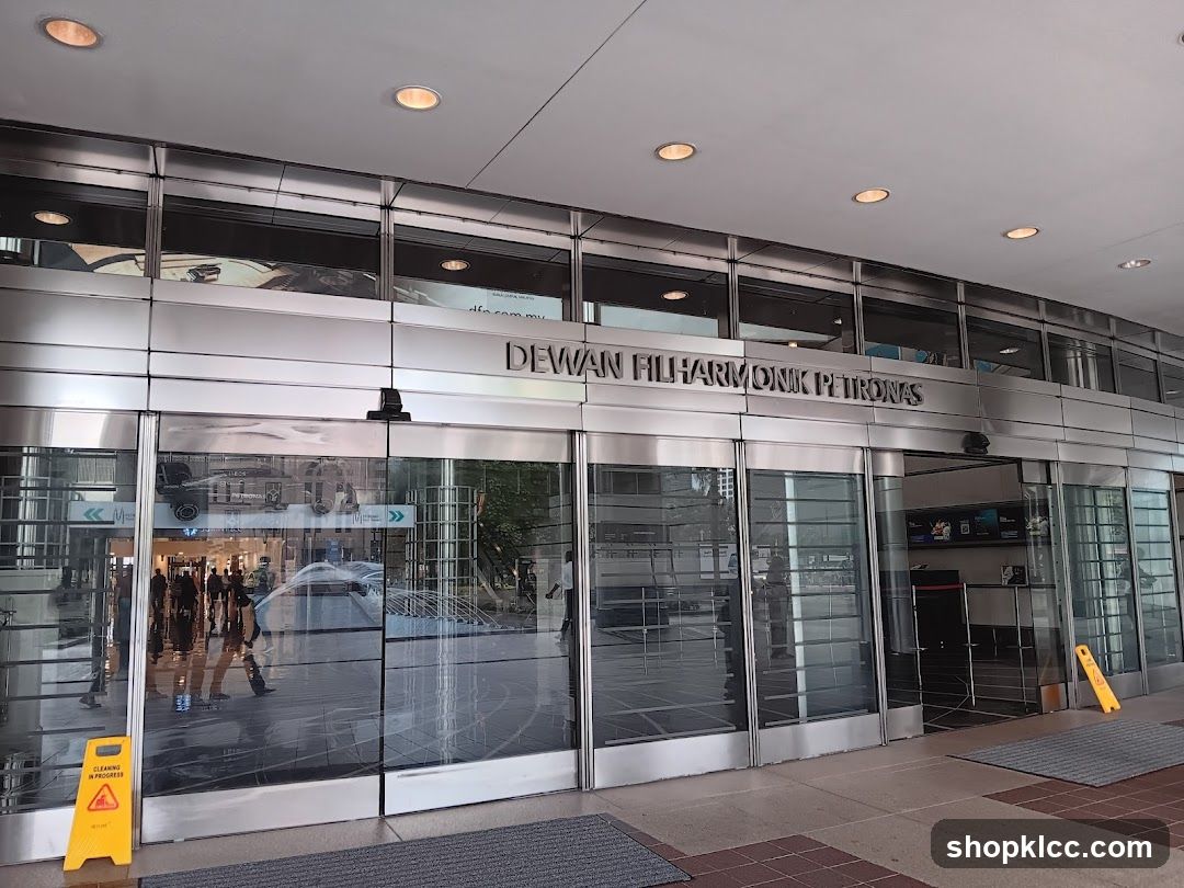 The exterior of the Dewan Filharmonik Petronas building features large glass doors and windows, with the name of the establishment prominently displayed above the entrance. The facade is modern and sleek, with metallic accents. Inside, people can be seen walking through the lobby. A yellow "Cleaning in Progress" sign is visible near the entrance.