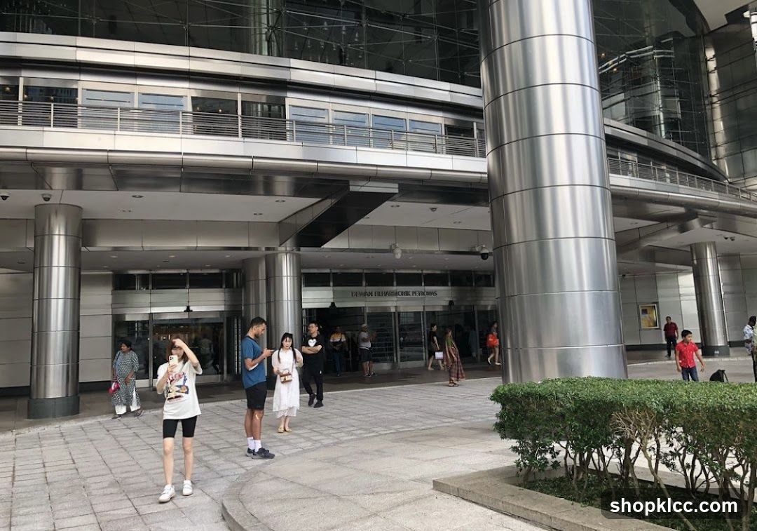 A wide shot of the entrance to Dewan Filharmonik Petronas, a concert hall. People are seen walking in and out of the building. The building has a modern architectural design with large glass windows and metallic columns.