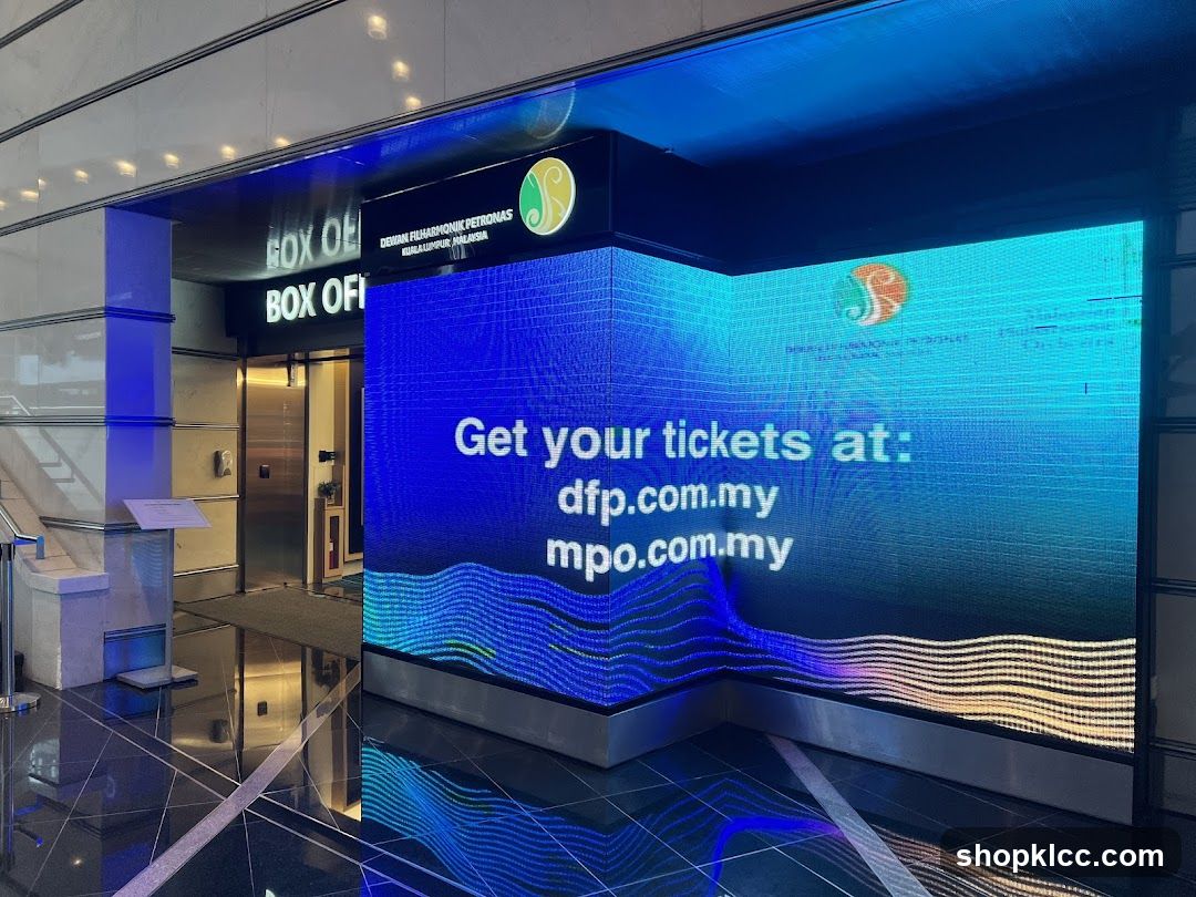 The entrance to a box office with a sign that reads "BOX OFI" and "BOX OF". Above the entrance, a sign displays "DEWAN FILHARMONIK PETRONAS KUALA LUMPUR MALAYSIA" and a logo. To the right, a large LED screen displays "Get your tickets at: dfp.com.my mpo.com.my" with wavy blue and yellow graphics.