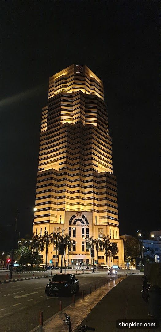 A tall, illuminated building at night with the name "PUBLIOBANK" visible above the entrance. Palm trees line the front of the building, and a street with cars is in the foreground.
