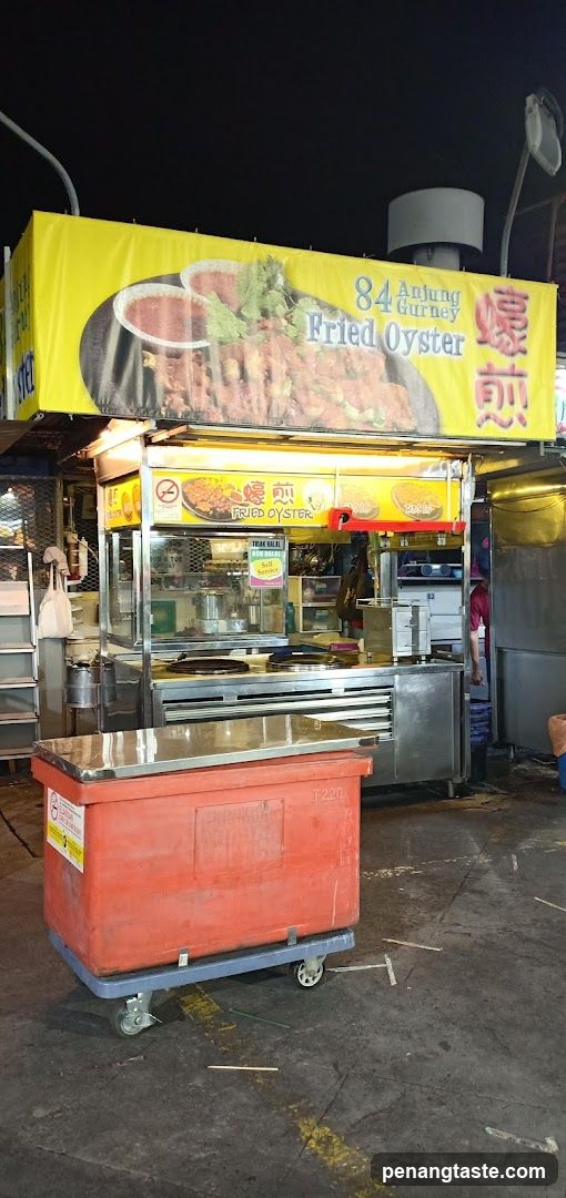 A food stall at night with a large yellow banner displaying "84 Anjung Gurney Fried Oyster" and an image of fried oysters. Below the banner is the stall itself, with glass enclosures and cooking equipment. An orange cooler on wheels is in the foreground.