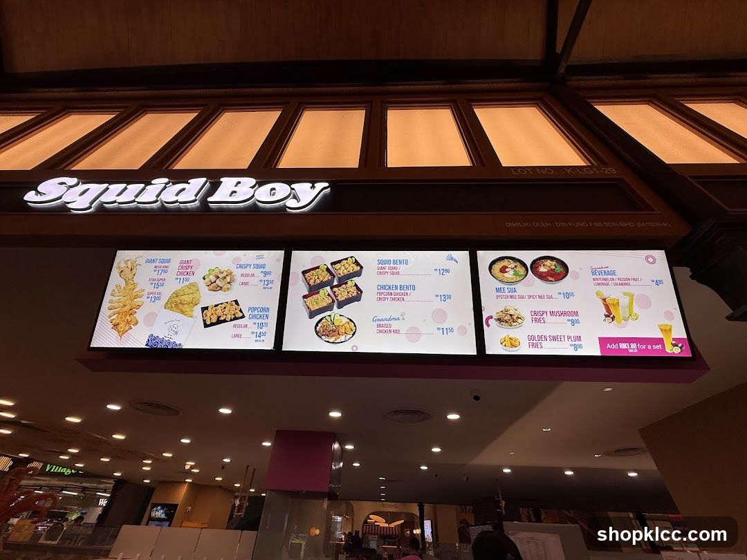 A digital menu board displaying food items and prices from Squid Boy, featuring bento boxes, fried chicken, mee gao, and desserts.