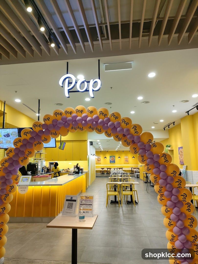 The image shows the entrance to a store called "Pop". The entrance is decorated with a balloon arch in yellow and purple. Inside the store, there is a yellow counter with staff members, and seating areas with yellow chairs and tables. The walls are painted yellow, and there are some posters on the walls.