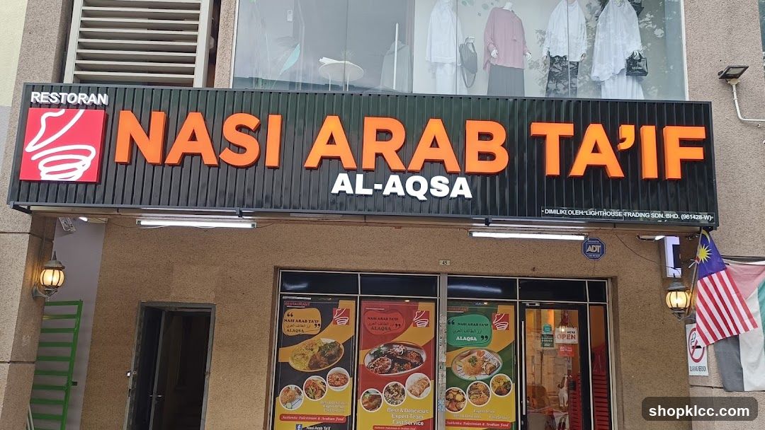 The exterior of a restaurant with a large sign that reads "RESTORAN NASI ARAB TA'IF AL-AQSA". Below the sign are three large posters displaying various dishes. The entrance to the restaurant is visible, with a sign indicating it is open. A Malaysian flag is also displayed.