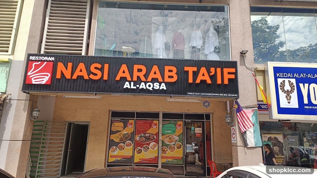 The storefront of a restaurant named Nasi Arab Ta'if Al-Aqsa. The restaurant has a large black sign with orange lettering that reads "RESTORAN NASI ARAB TA'IF AL-AQSA". Below the sign, there are three large posters displaying various dishes. The entrance to the restaurant is visible on the left side of the image. To the right of the restaurant, there is a sign for "KEDAI ALAT-ALA YO".