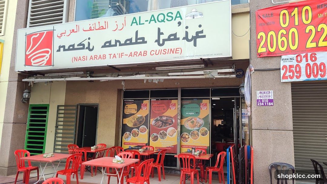 The exterior of Al-Aqsa restaurant, a shop front with a large sign displaying the restaurant name in Arabic and English, along with images of food. Red tables and chairs are set up outside the restaurant. Promotional posters are displayed on the windows.