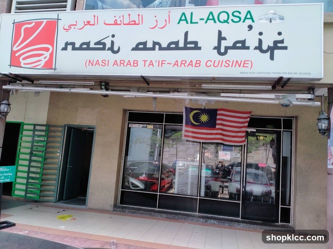 The exterior of a restaurant with a large sign above the entrance. The sign displays the restaurant name in Arabic and English, "AL-AQSA" and "(NASI ARAB TA'IF~ARAB CUISINE)". A Malaysian flag is displayed on the storefront window. The entrance has large glass windows and doors. To the left of the entrance, there are green metal racks and an open doorway.