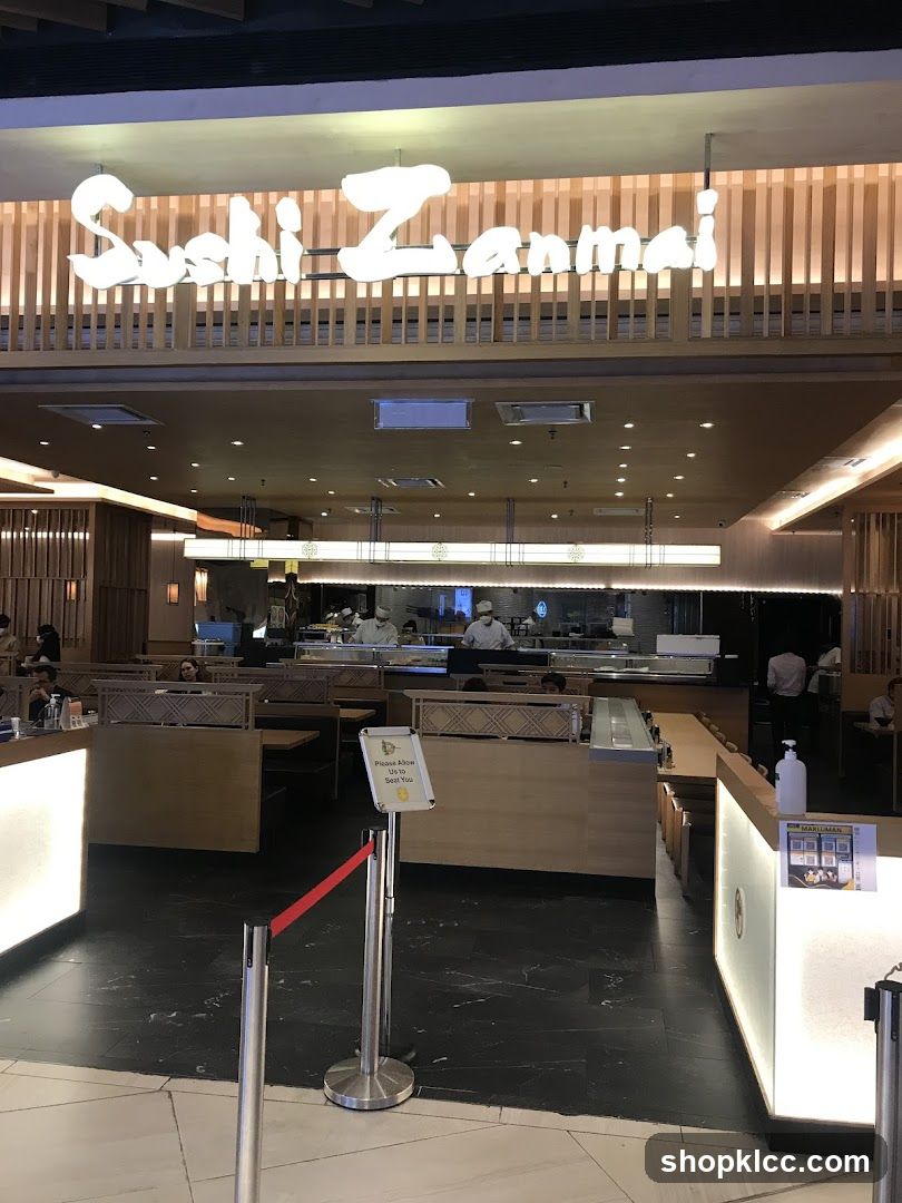 The exterior of a Sushi Zanmai restaurant. The large, illuminated sign with the restaurant's name is prominently displayed above the entrance. Inside, the restaurant appears to have a sushi bar and seating areas. Staff members in chef uniforms are visible behind the sushi bar.