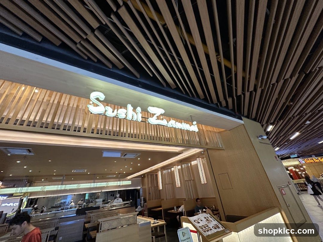 The exterior of a Sushi Zanmai restaurant with a prominent illuminated sign. The interior is visible, showing a sushi bar and dining area with staff preparing food. The ceiling features a distinctive wooden slat design.
