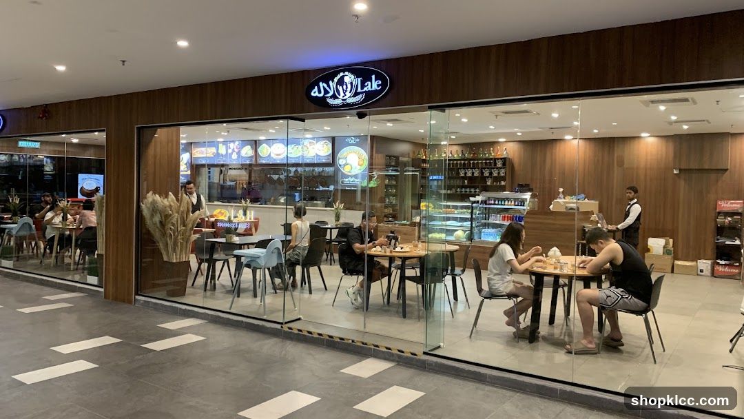 A wide shot of the exterior of a restaurant called Lale, with large glass windows and a wooden facade. Inside, several patrons are seated at tables, and staff members are visible. The restaurant has a modern interior with display cases and shelving.