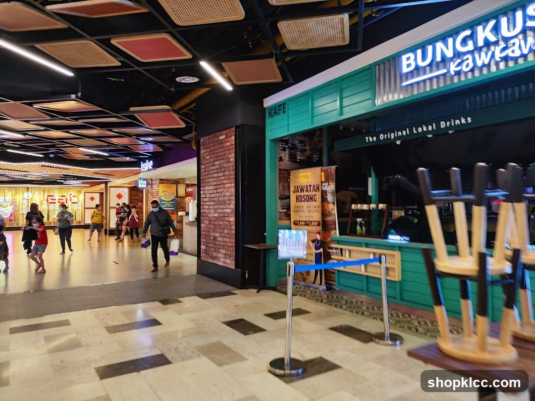 A teal-colored shop front with neon signage that reads "BUNGKUS KAWKAW" and "The Original Lokal Drinks". There is a job posting for "JUNIOR KITCHEN BARISTA HELPER" with a salary range of 1,800-2,800. Stools are stacked on tables in front of the counter.