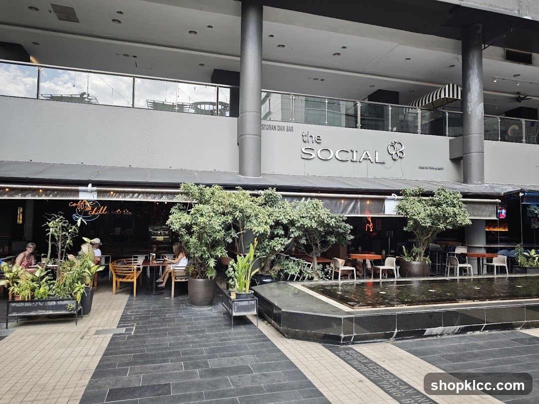 The exterior of a restaurant and bar called "The Social" is visible. The establishment has outdoor seating areas with tables and chairs, surrounded by potted plants and a water feature in the foreground. The storefront features large windows and a prominent sign with the restaurant's name.