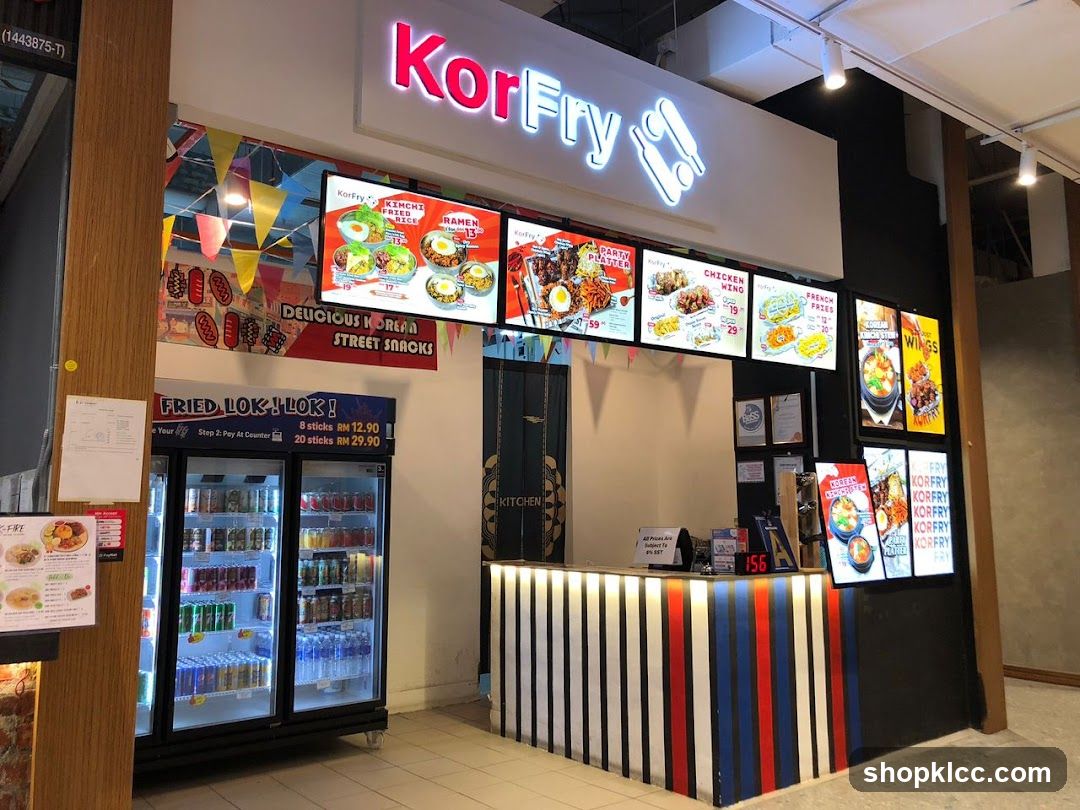 The exterior of a Korean street food stall named KorFry. Digital menu boards display various Korean dishes like Kimchi Fried Rice, Ramen, Chicken Wings, and Party Platters. A refrigerator stocked with drinks is visible, along with a counter displaying prices for "Fried Lok! Lok!".