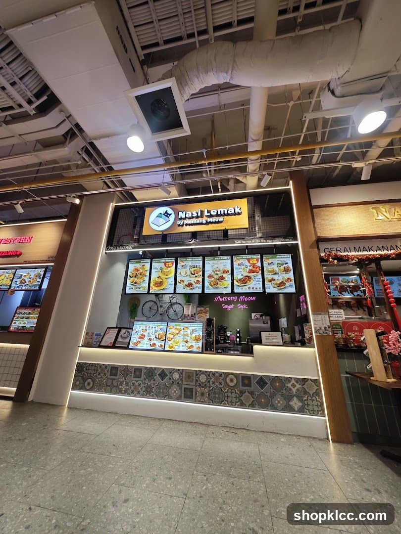 A food stall with a prominent sign displaying "Nasi Lemak by Memang Meow" and a cartoon cat logo. The stall features digital menu boards showcasing various dishes, a decorative bicycle, and a counter area. The lower part of the stall is adorned with patterned tiles.