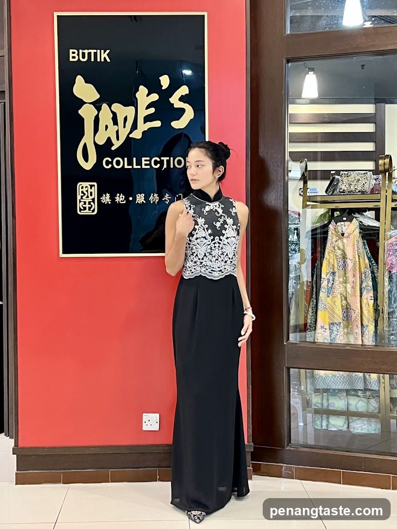 A woman stands in front of a red wall with a large black sign that reads "BUTIK Jade's COLLECTION" in gold lettering. To the right, a glass display window shows racks of colorful garments.