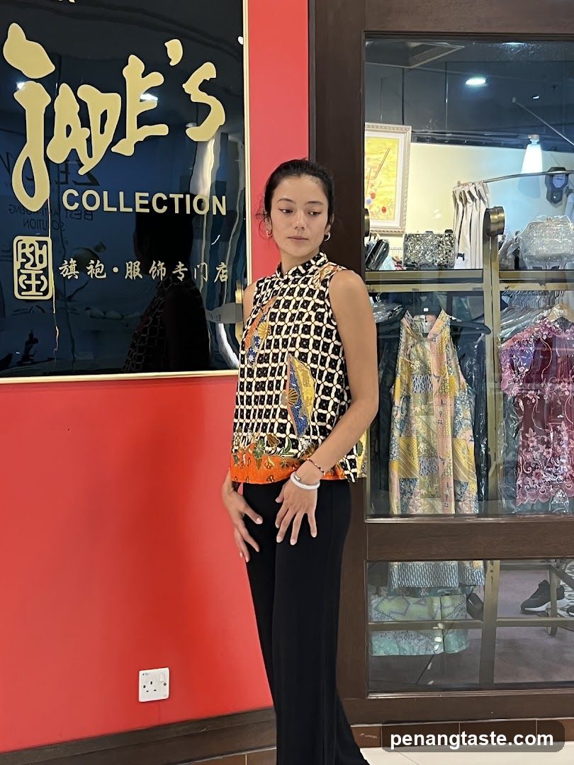 A woman stands in front of a shop with a red wall and a large black sign that says "Jade's Collection" in gold lettering. The shop window displays various clothing items and accessories.