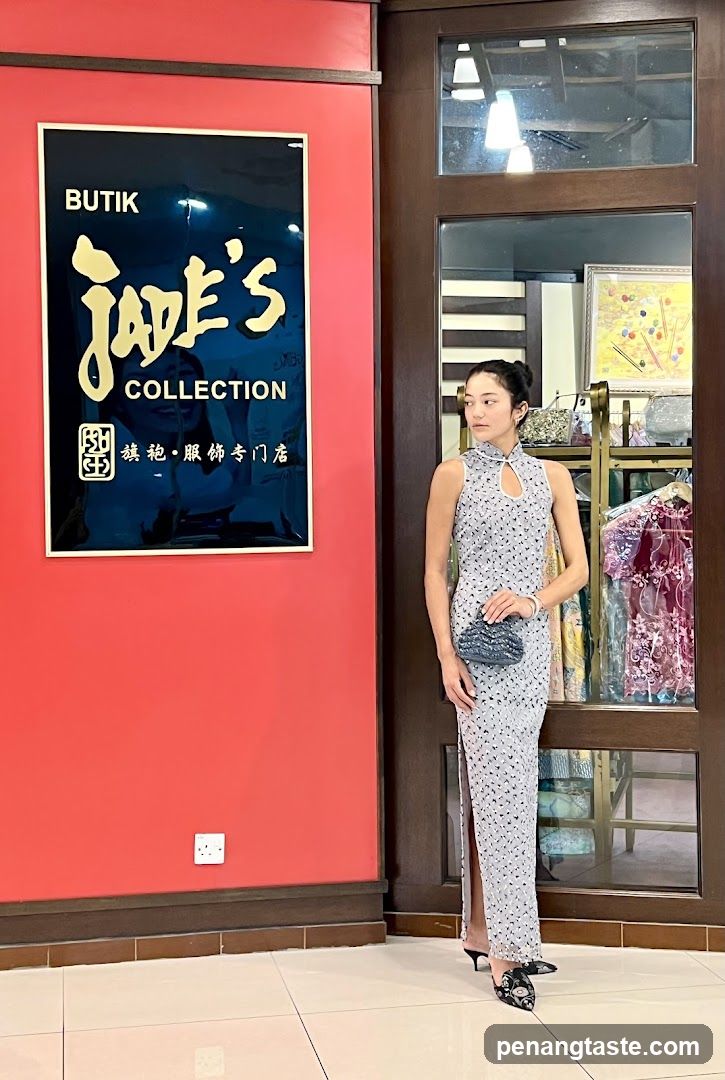 A woman in a patterned dress stands in front of a red wall with a sign that reads "BUTIK Jade's COLLECTION". The sign is black with gold lettering. To the right of the woman is a glass-paned door, through which clothing racks and displays can be seen.