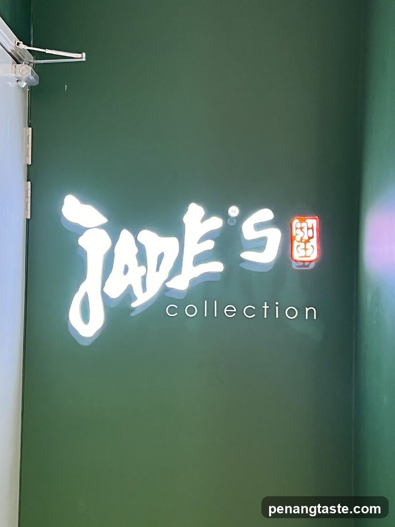 A white neon sign with the words "Jade's collection" is mounted on a dark green wall. The sign has a stylized font for "Jade's" and a simpler font for "collection". To the right of "Jade's" is a small, illuminated red square with a symbol inside. A door hinge and part of a white door are visible on the left side of the image.