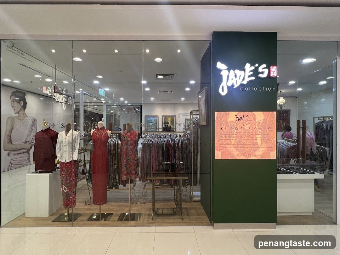 The storefront of Jade's Collection features a large glass window displaying mannequins dressed in traditional attire, including cheongsams and patterned skirts. The shop's name is prominently displayed in illuminated white lettering on a dark green pillar. Below the name, a framed document with Chinese characters and English text provides information about the collection. Racks of clothing are visible inside the store, along with display shelves and a vintage sewing machine.