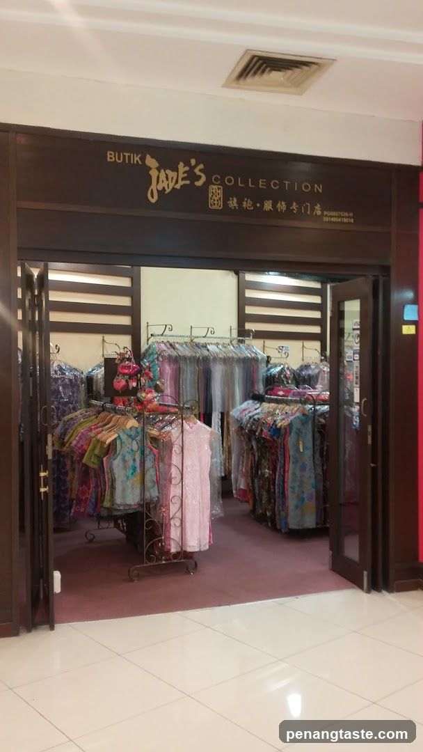 The exterior of a clothing boutique with the name "BUTIK JADE'S COLLECTION" displayed above the entrance. The store features racks of colorful clothing, including dresses and tops, visible through open doors.