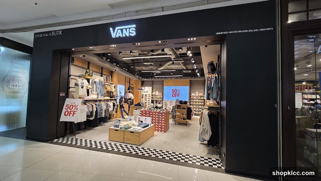 The exterior of a Vans store with the brand's logo prominently displayed above the entrance. Inside, clothing and shoes are displayed on shelves and racks. A large screen shows a "50% OFF" promotion. Signs indicating "UP TO 50% OFF" are also visible outside the store.