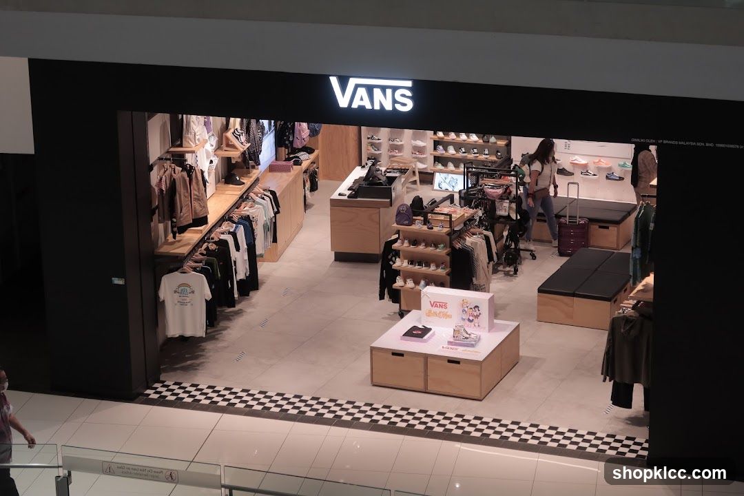 The exterior of a Vans store with the Vans logo prominently displayed above the entrance. Inside, clothing and shoes are displayed on shelves and racks. Several customers are browsing the merchandise.
