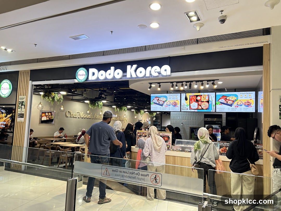 The exterior of a Dodo Korea restaurant with a prominent sign above the entrance. The restaurant features a modern design with wooden accents and hanging plants. Digital menu boards display images of food items and prices. Several customers are visible inside and queuing outside.
